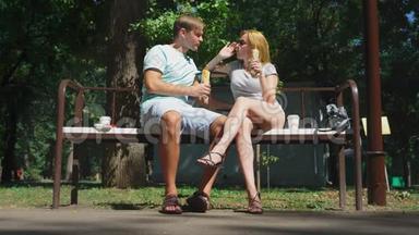 快乐的一对夫妇<strong>吃热狗</strong>，在城市公园的长凳上玩得很开心。 4K，慢动作。